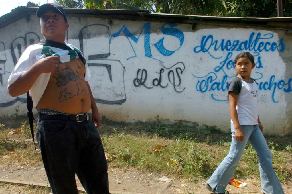 MS-13 member shows off his gang tattoos in front of a wall covered in gang graffiti.