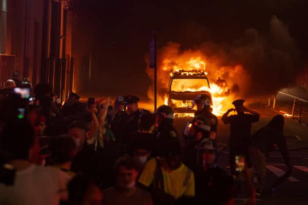 Protesters in front of a police car that has been set one fire during a protest